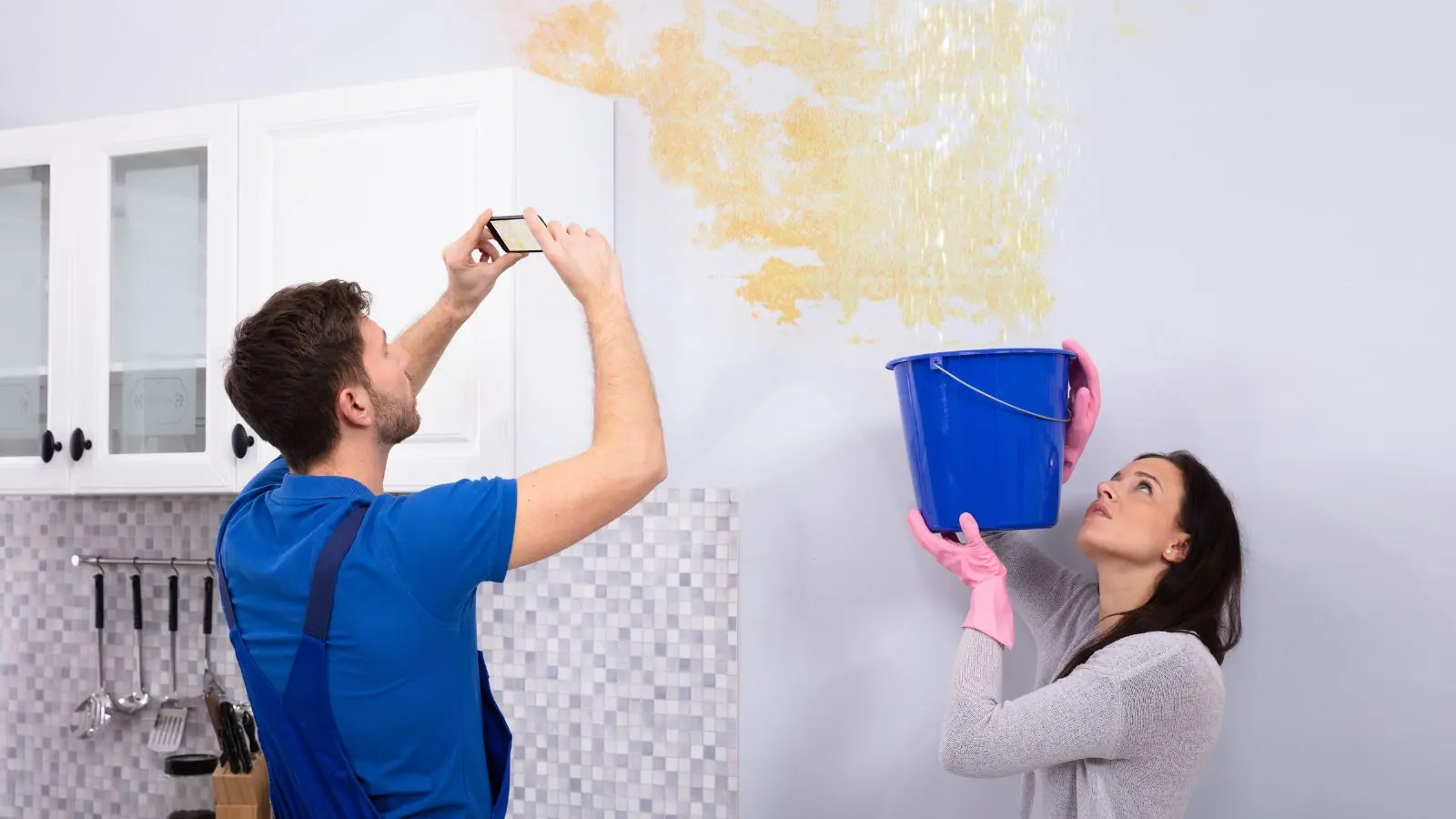 Homeowner photographing a stained wall while a bucket catches a leak